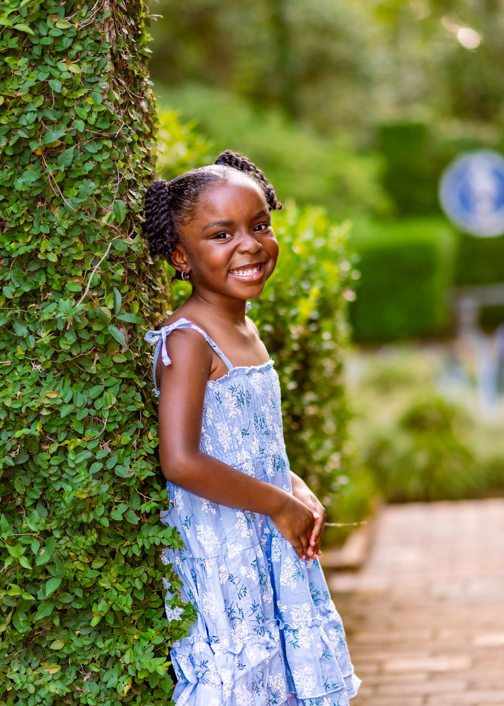 A smiling young Black girl in a blue floral dress leans against an ivy-covered tree in a lush green garden.
