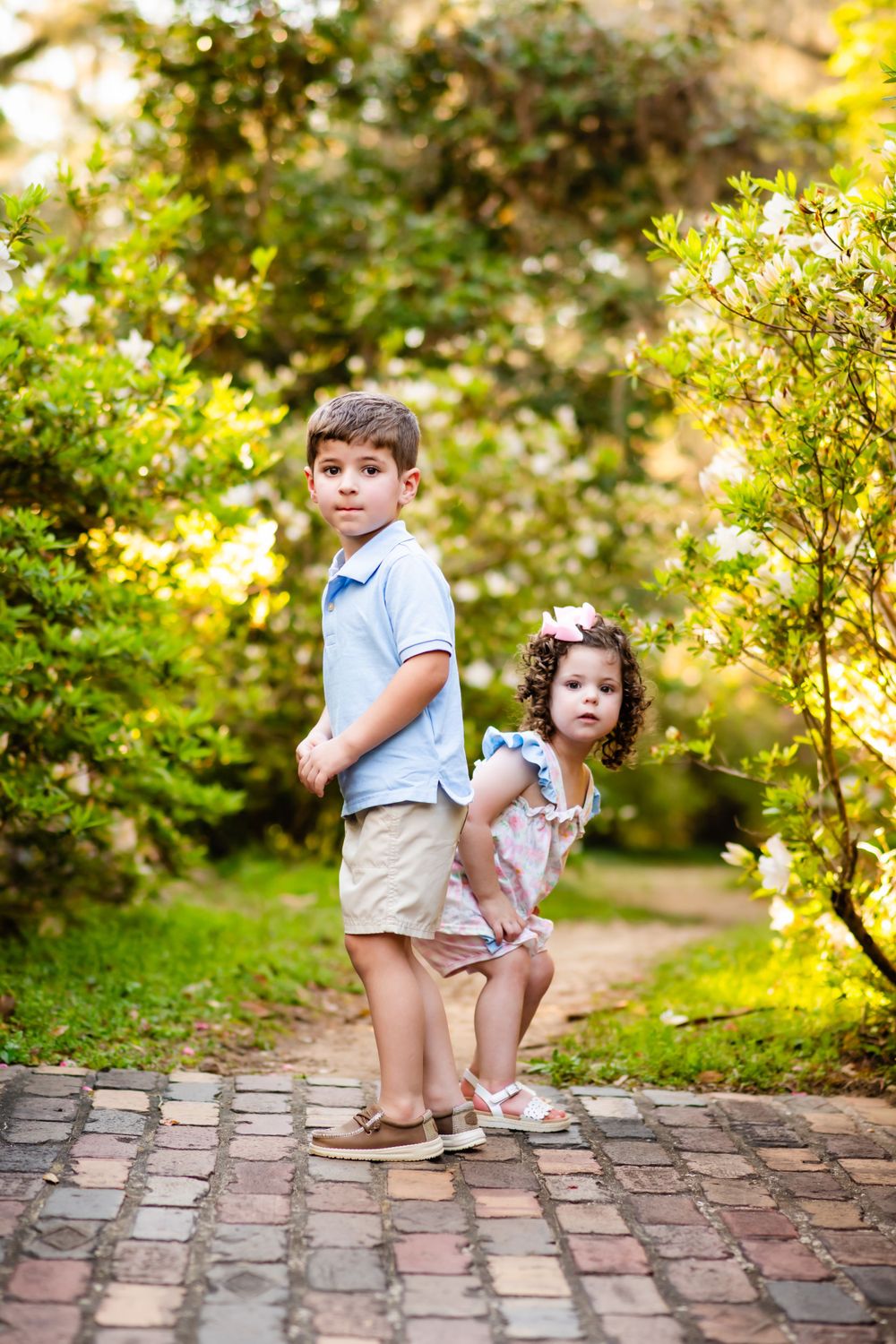 Two children pose playfully on a brick pathway surrounded by lush greenery and flowers in a garden setting.