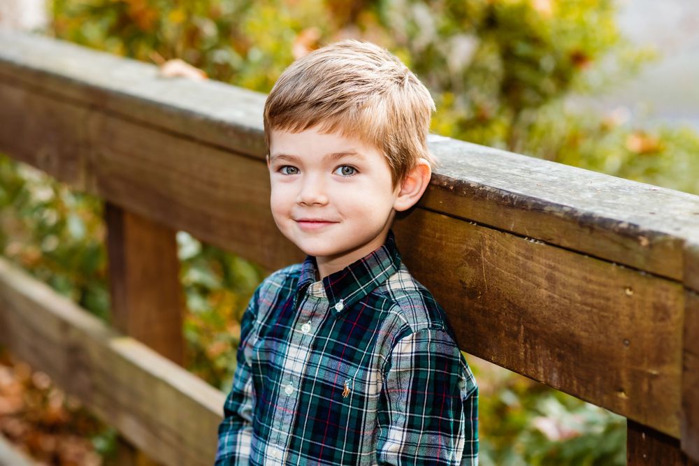A young person in a plaid button-down shirt poses outdoors against a rustic wooden railing in autumn.