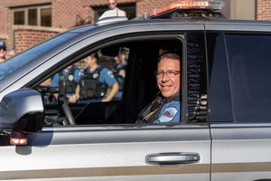 A law enforcement officer smiles while sitting in a police vehicle parked outside a brick building.