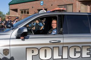 A police officer smiles while sitting in an official police vehicle outside a brick building.