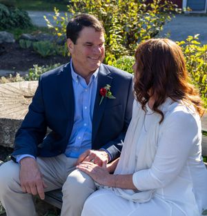 A couple in wedding attire sits together outdoors, sharing a sweet moment on a sunny day.