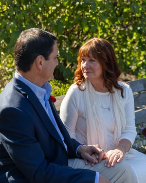 A couple in formal attire sits together on a bench outdoors surrounded by green foliage in the background.