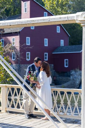 A couple shares a romantic moment on a white wooden bridge railing with a rustic red building in the background.