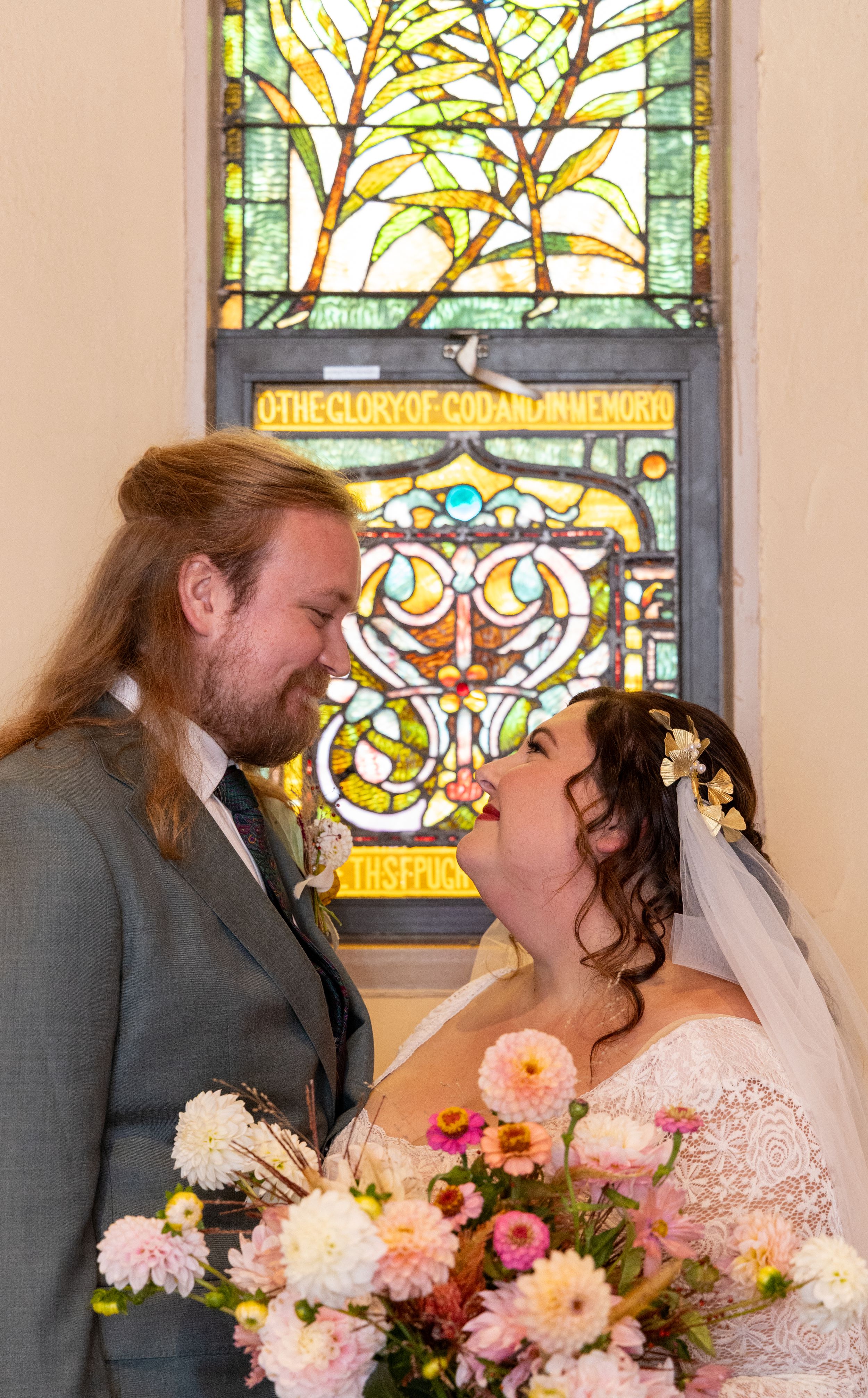 A couple shares a romantic moment in front of a beautiful stained glass window while holding a pink floral bouquet.