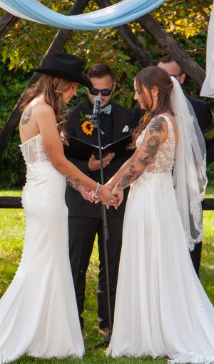 Wedding ceremony under blue draping with two brides in white gowns and sunflower boutonniere.