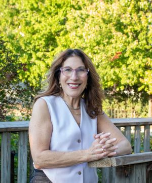 Professional portrait of woman in white vest standing by wooden railing with greenery background.