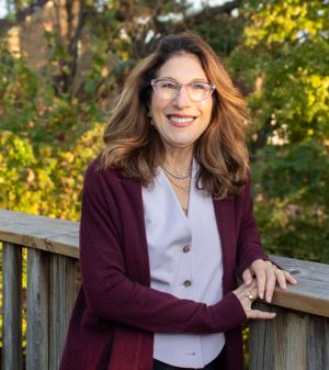 Professional headshot of woman in burgundy cardigan and white top by wooden railing outdoors.
