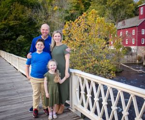 Family portrait on white wooden bridge with red mill building in autumn background.