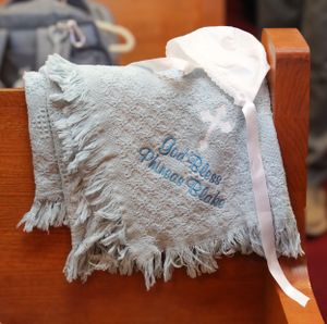 A white decorative baby pillow with blue embroidery and fringe detail resting on wooden furniture.