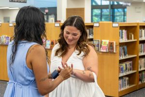 Two women with tattoos chat and laugh together inside a library with bookshelves in the background.