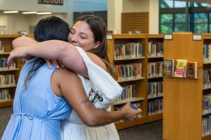 Two women sharing a warm hug in a library, wearing blue and white dresses among bookshelves.