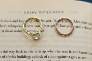 Two gold rings resting on an open Casey McQuiston book, one featuring a diamond solitaire setting.