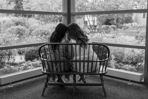 Two girls sit close together on a wooden bench by a large window overlooking a lush garden in black and white.