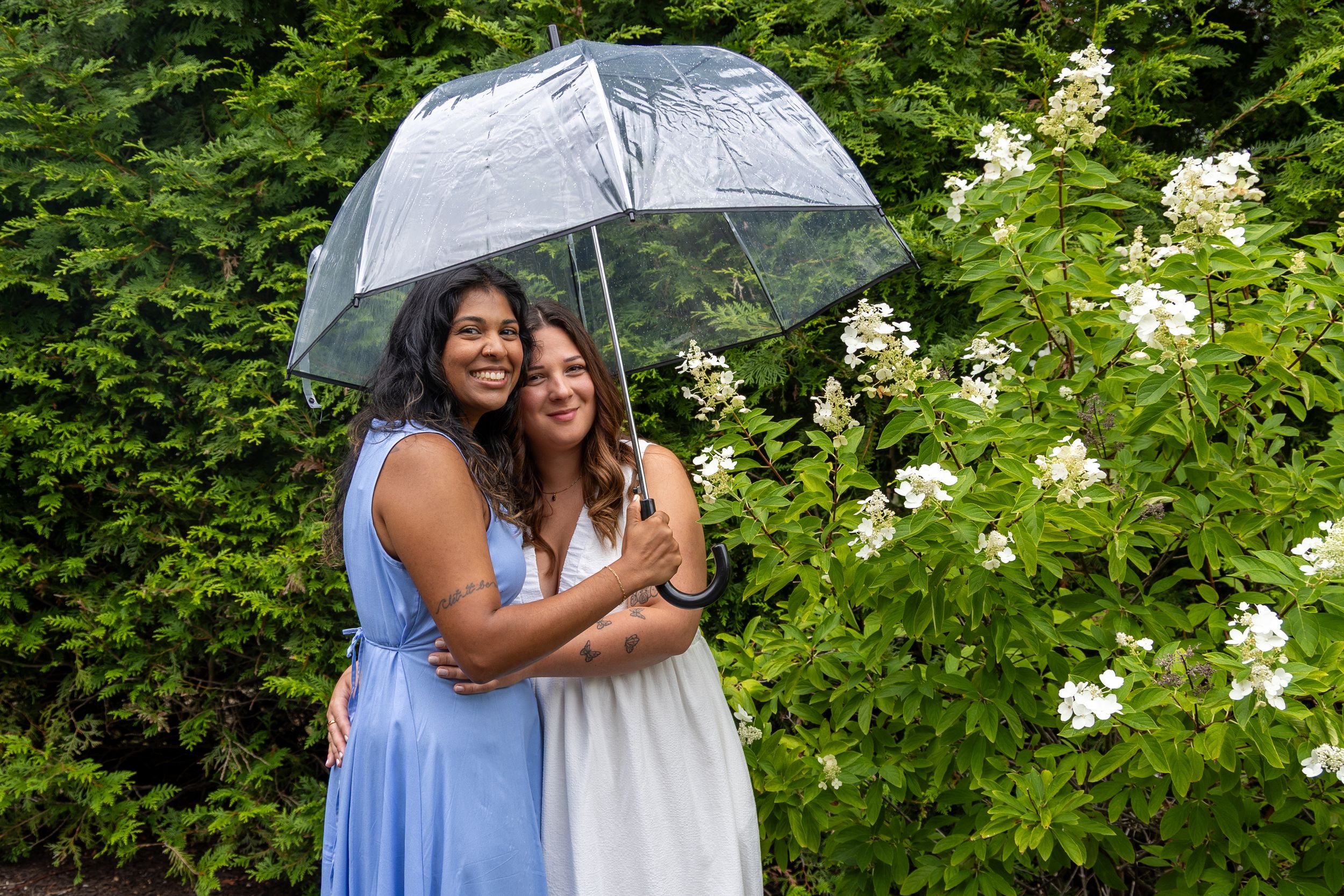 Two women smiling under a clear umbrella in a garden with white flowers, one in blue dress, one in white.