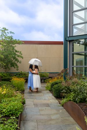 Two women sharing a white umbrella on a garden path, one in blue dress, one in white, near a modern building.