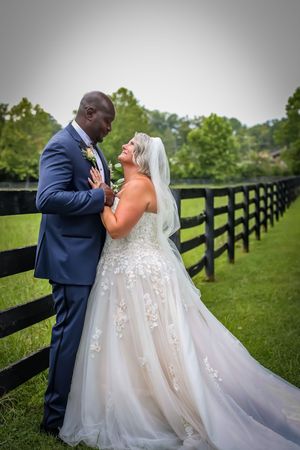 Newlywed couple shares an intimate moment along a rustic wooden fence line in a lush green field.