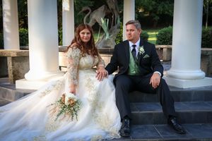 Wedding couple poses on classic white columned porch with elegant landscaping.