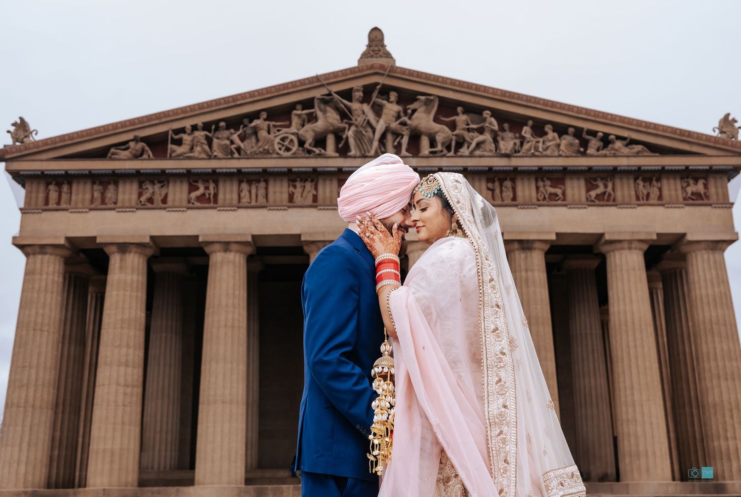 A bride in a pink and ivory gown and veil embraces her groom in a blue suit and pink turban before a grand columned Greek-style temple.