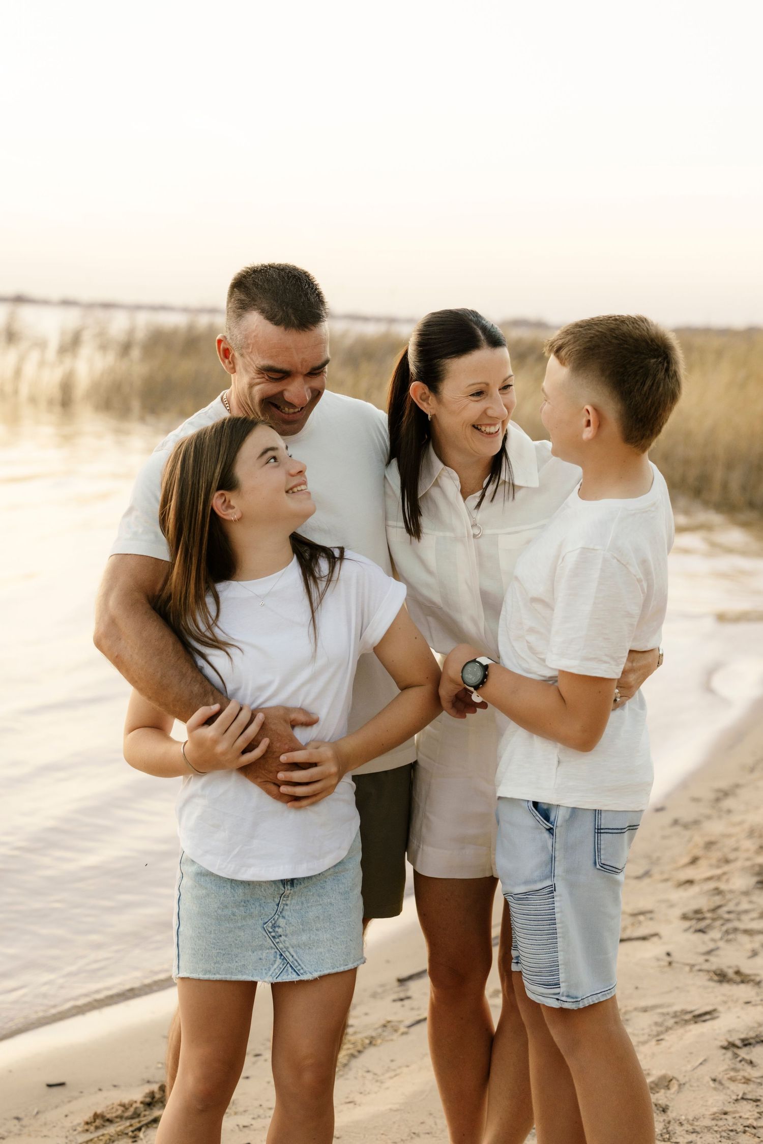 A happy family of four laughs together on a sandy beach at golden hour wearing white outfits.