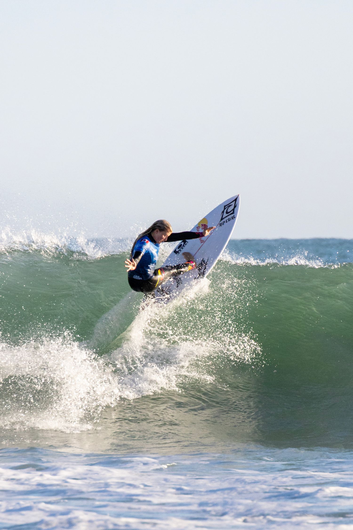 A surfer carves through a clean green wave beneath a clear coastal sky.