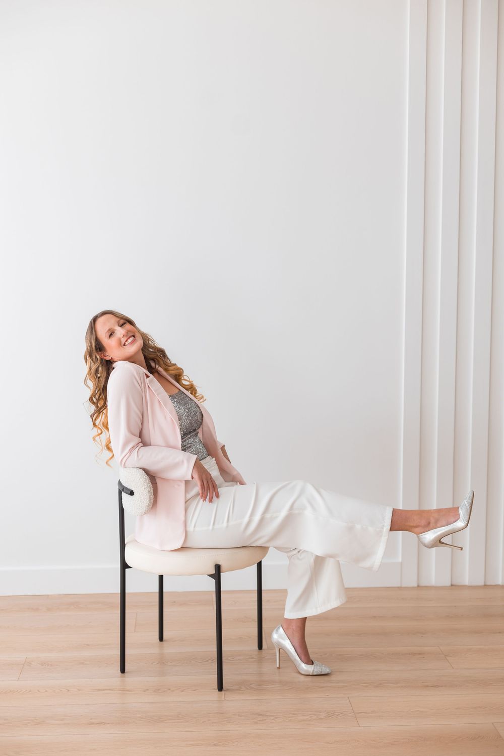 Smiling blonde woman in pink blazer and white pants sitting on chair against white wall.