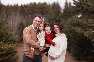 Family of four posing at a Christmas tree farm, parents holding two young girls, mother expecting, wearing festive holiday outfits.