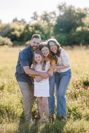 Happy family of four posing together in a sunny wildflower field, parents hugging two young daughters outdoors.