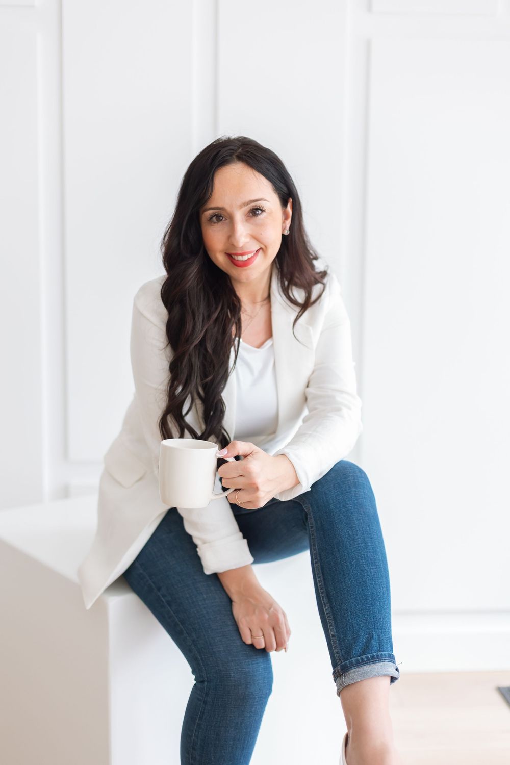 Confident woman in white blazer and jeans holding coffee mug against white wall.