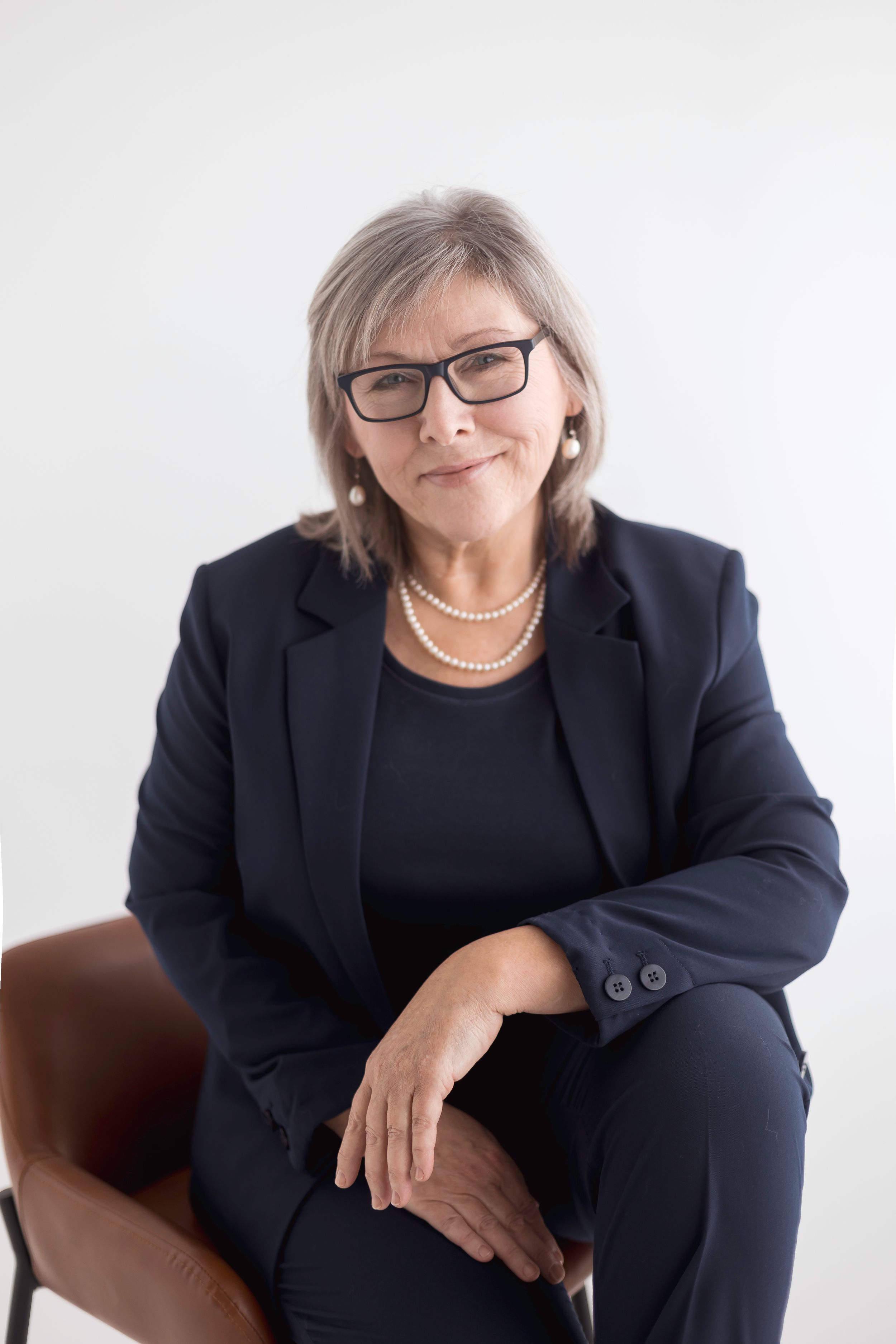 Senior businesswoman in navy suit with pearl necklace seated against white background.