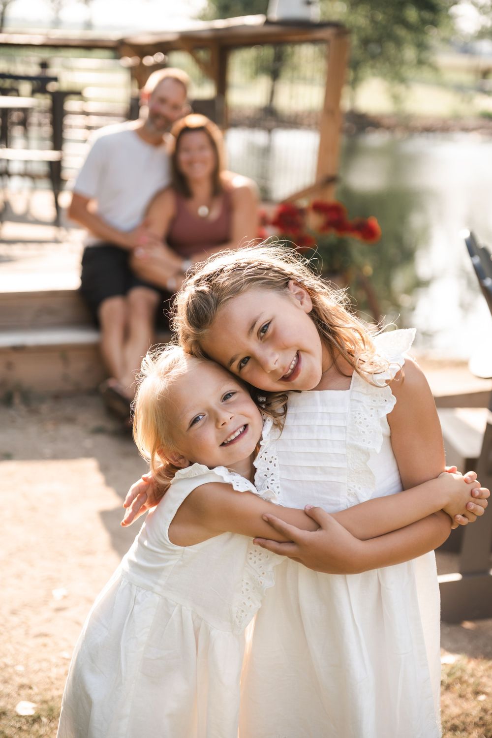 Two young children in matching white dresses embrace and laugh together in warm sunlight while adults sit in background.