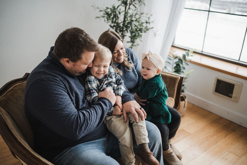 Family of four sitting together in a cozy chair, parents smiling at two young children in a bright, modern living room.