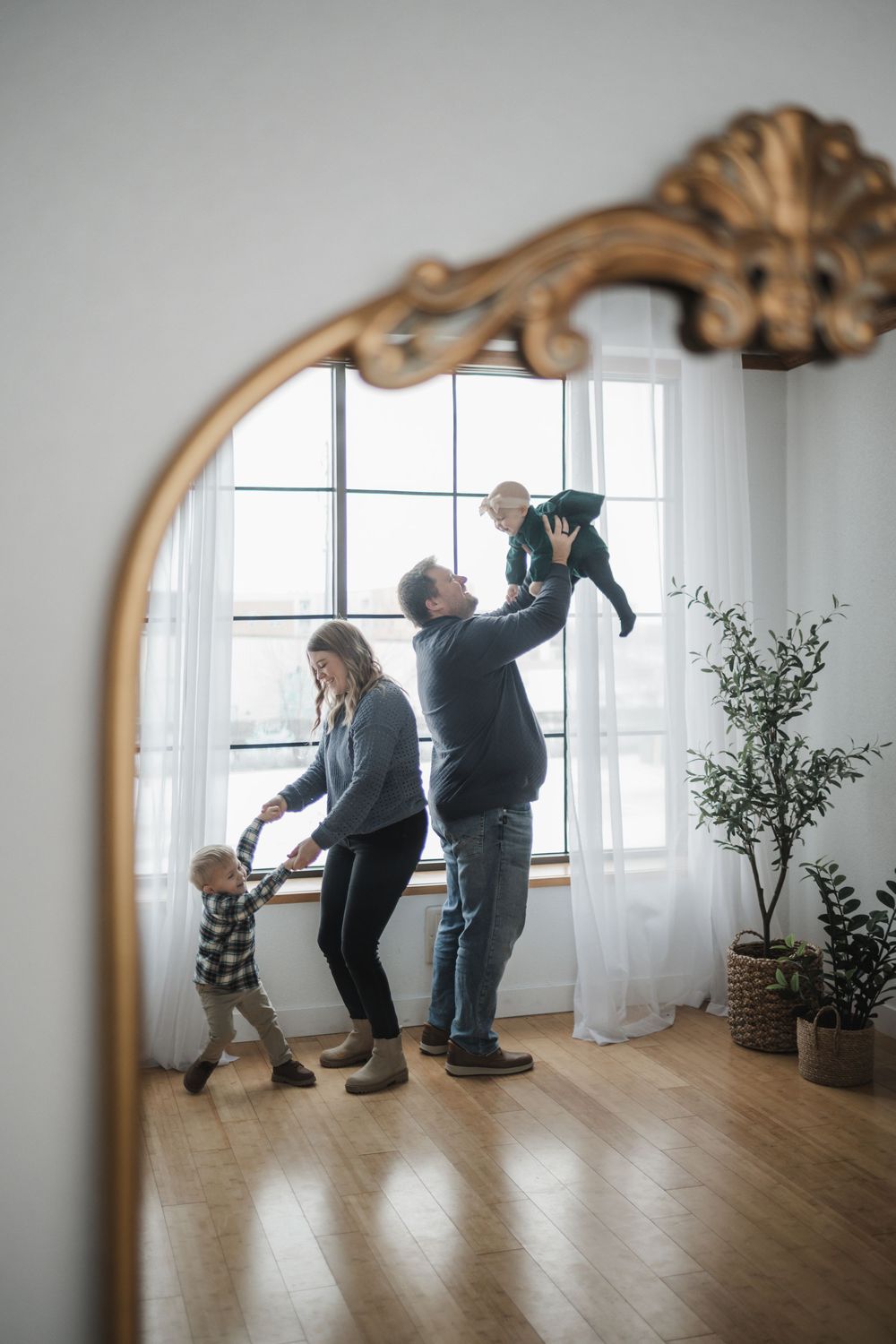 Family of four reflected in ornate gold mirror, parents playing with two young children in bright modern living room.