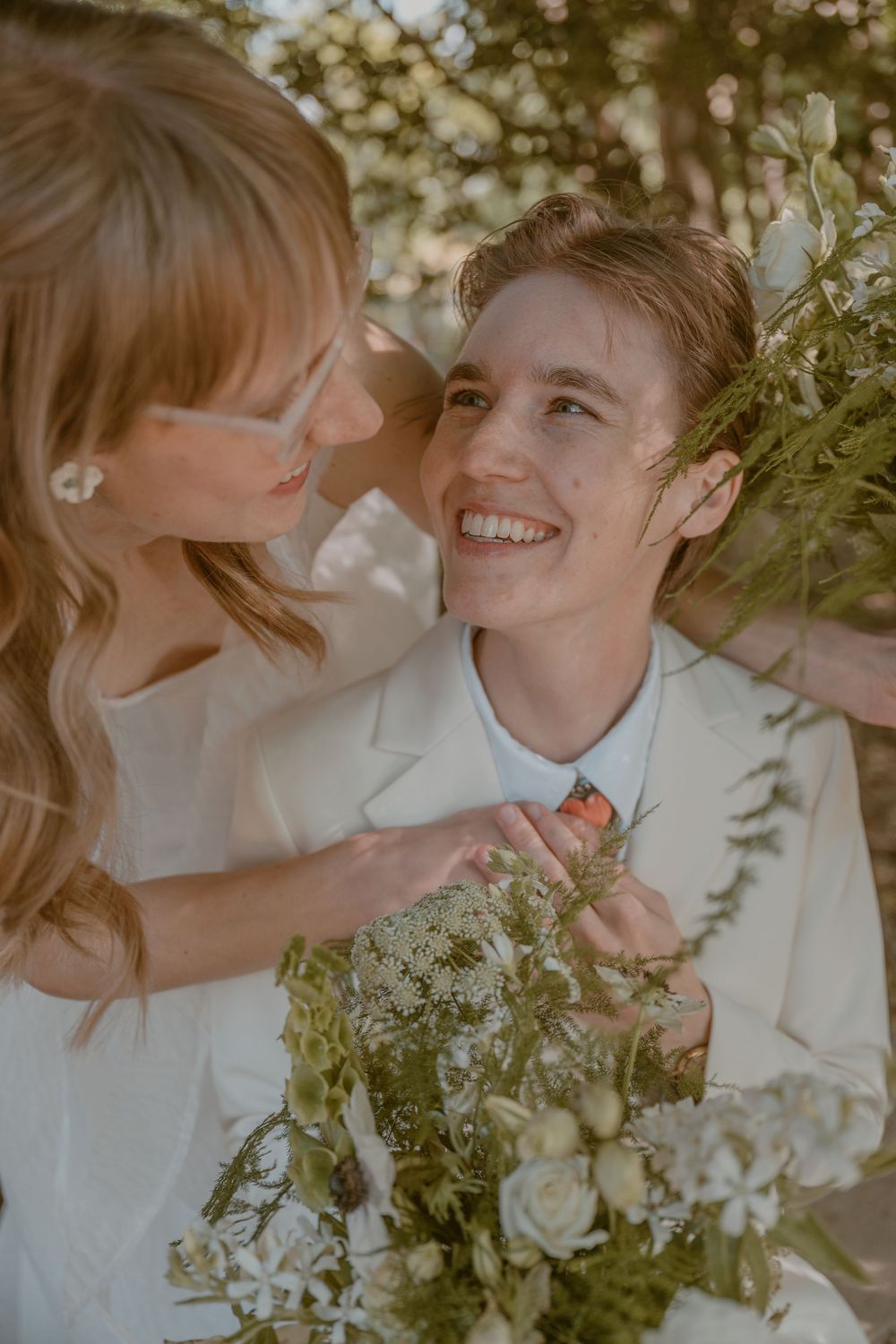 Two brides in white attire share a joyful moment, one smiling brightly while holding a wildflower bouquet outdoors.