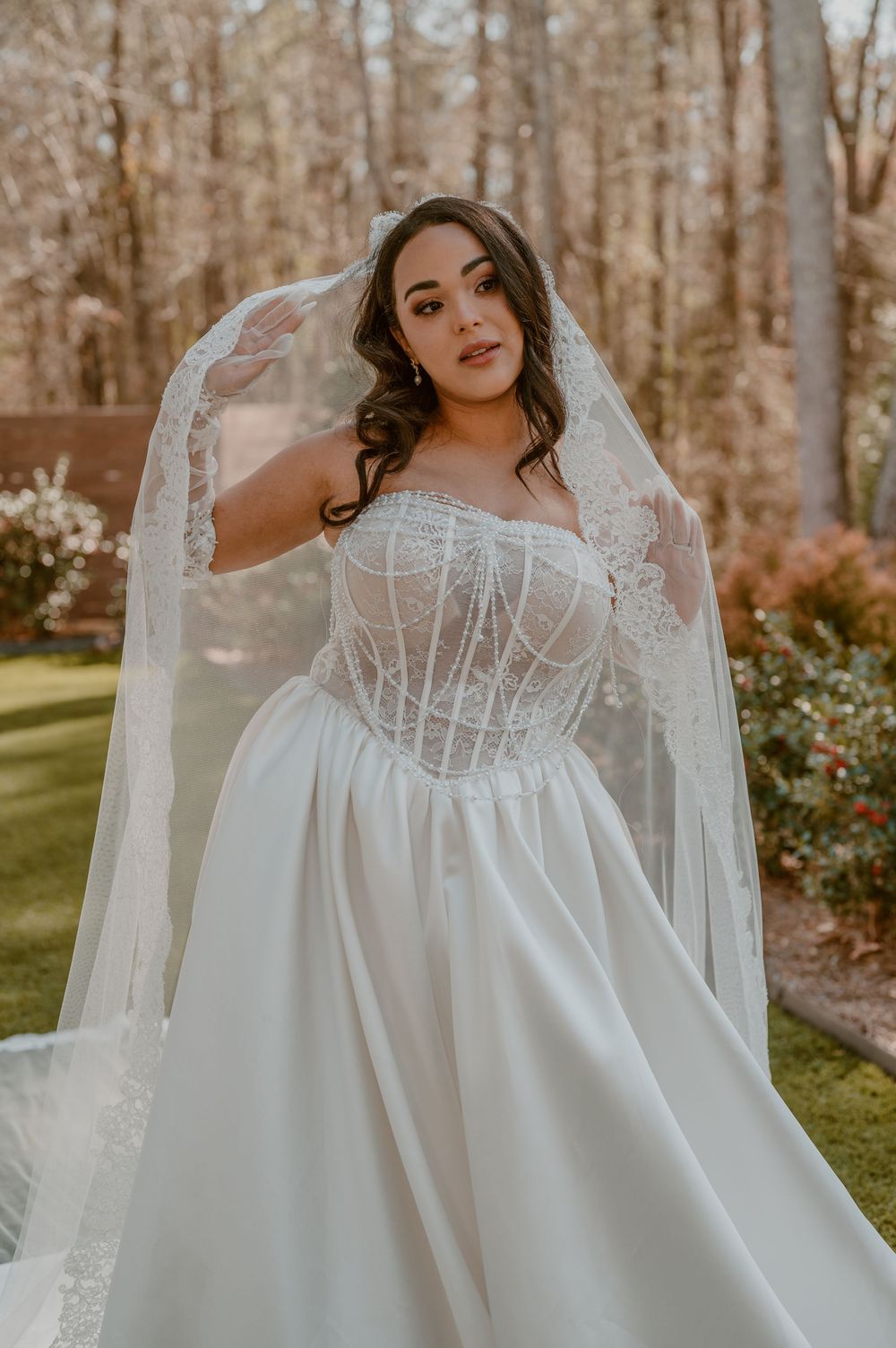 A bride poses in a white corset wedding gown with lace veil in a garden setting surrounded by autumn trees.