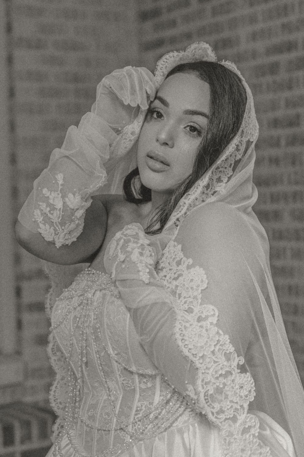 A black and white portrait of a bride wearing a delicate lace veil and long gloves against a brick wall.