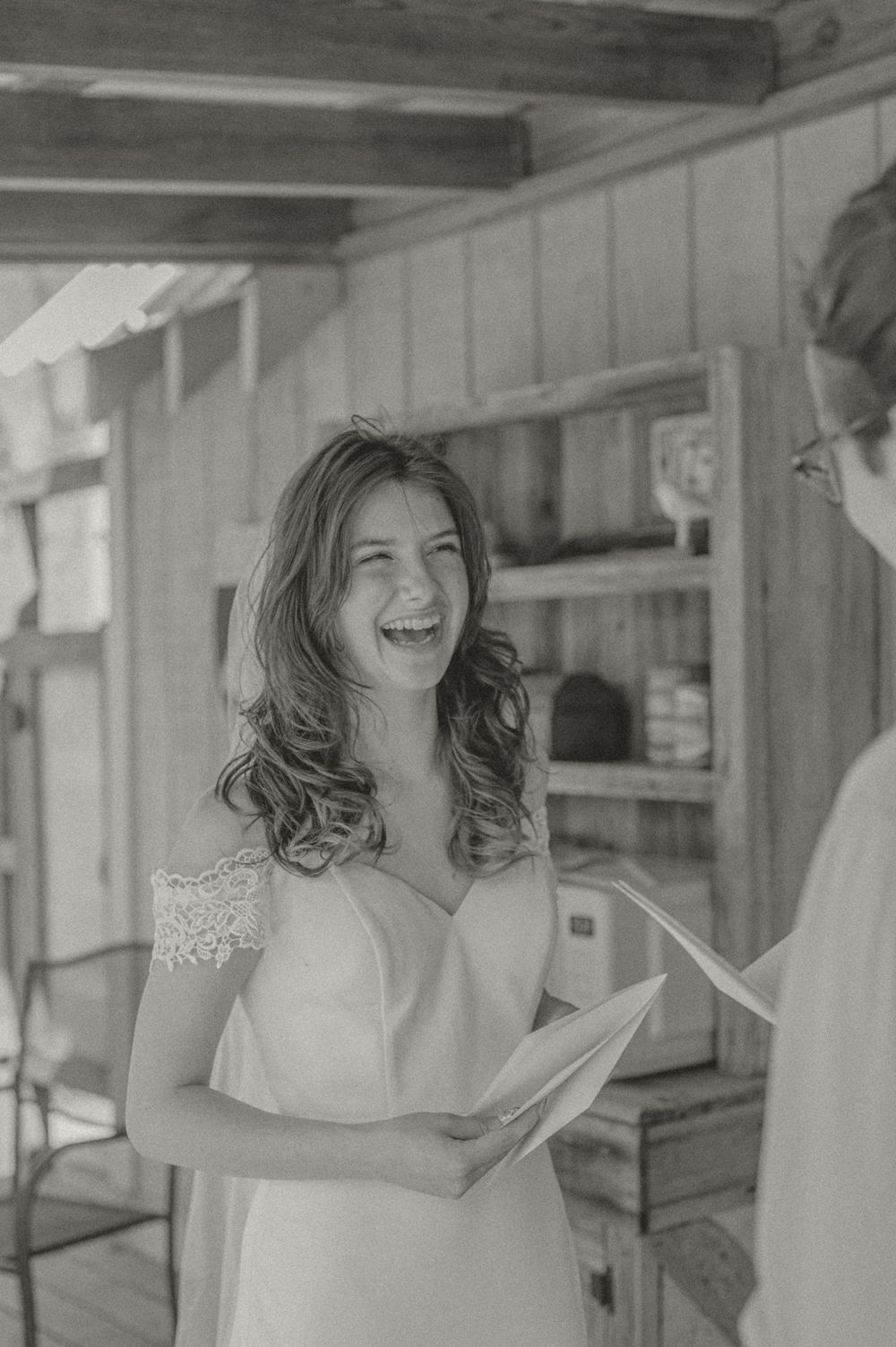 Black and white photo of a joyful bride laughing while holding papers in a rustic indoor setting.