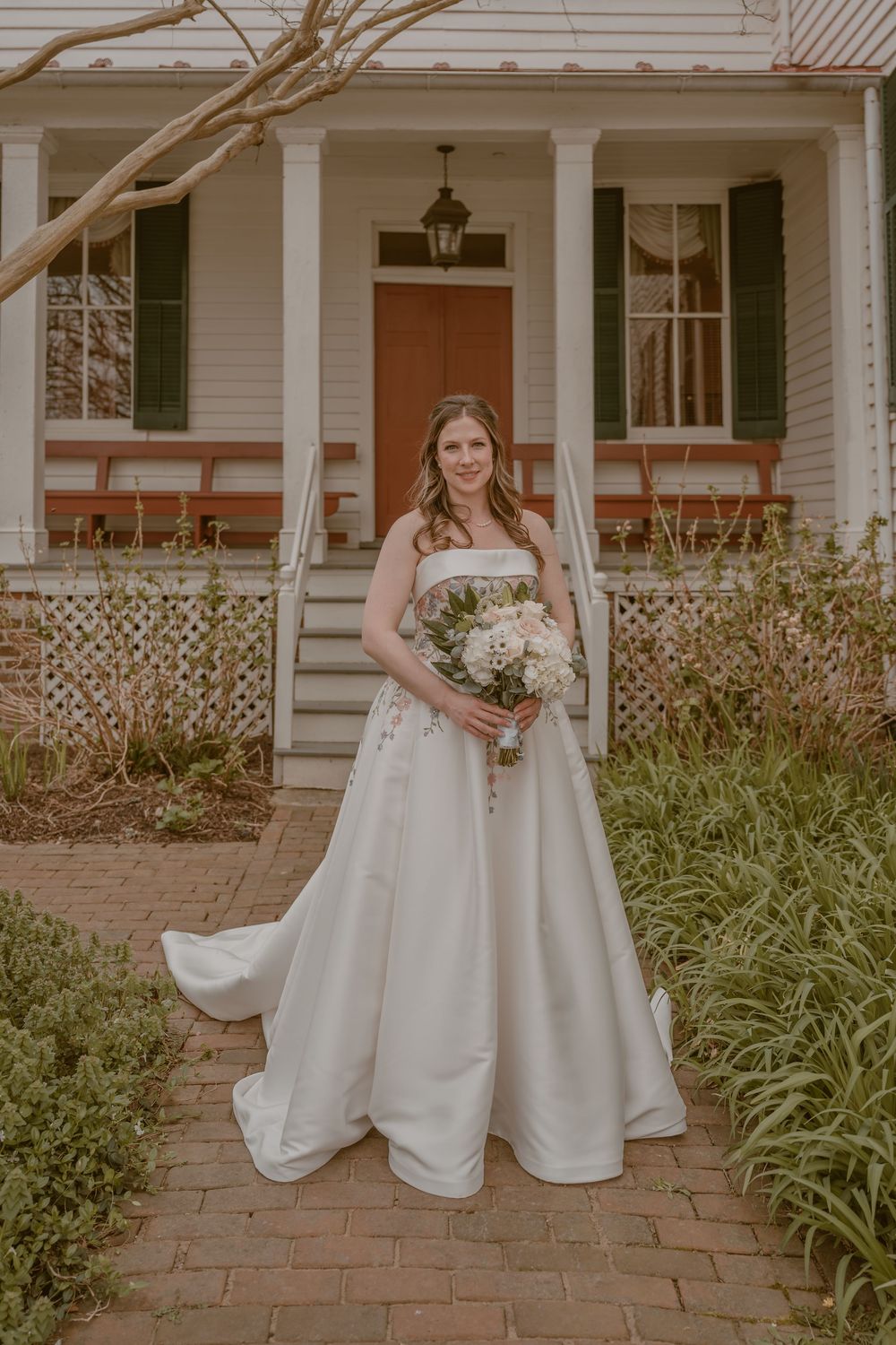 Bride in strapless white ball gown holding floral bouquet stands on brick path before charming white colonial home.