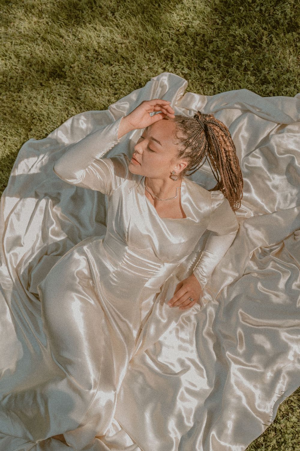 Person in flowing white satin dress laying on grass in dreamy sunlit outdoor setting.