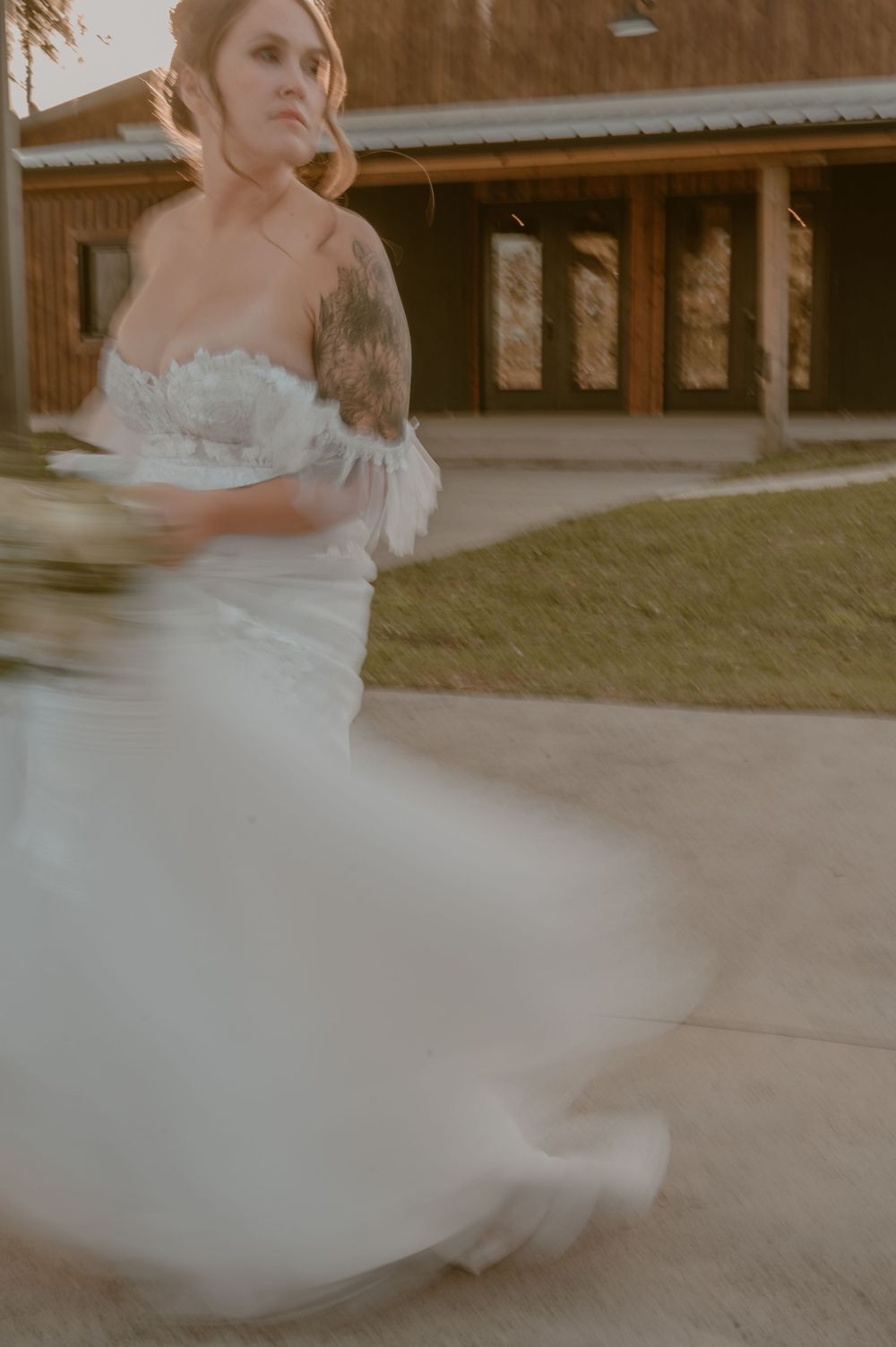 Ethereal motion blur captures a person in white wedding dress with tattoos twirling on patio near rustic wooden building.