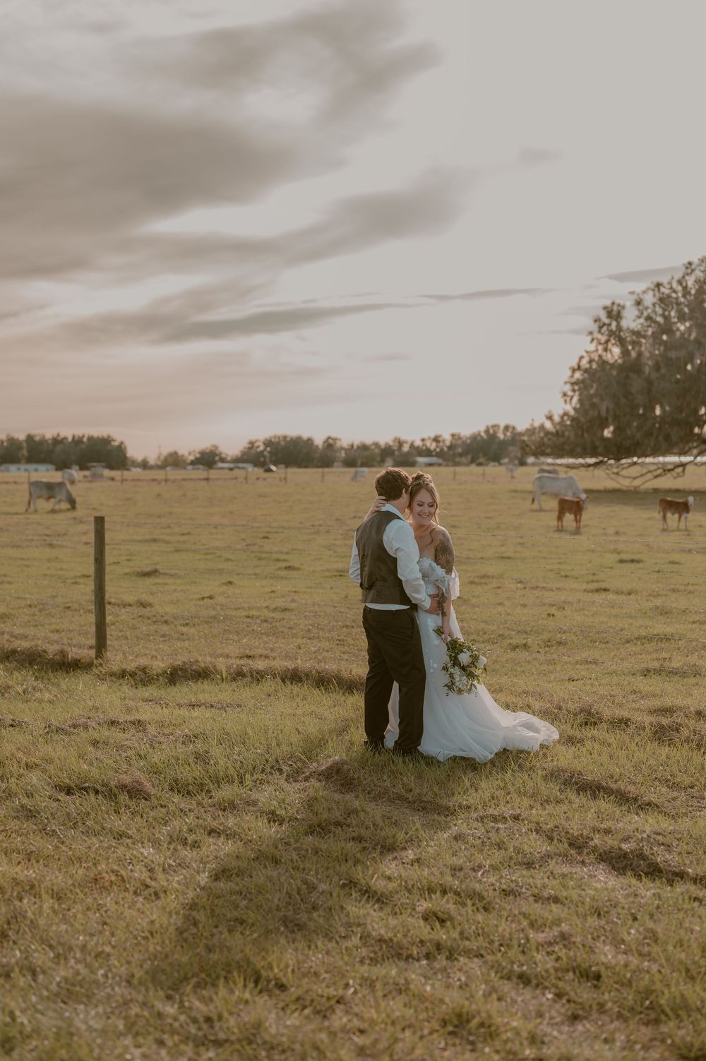 A couple embraces in a serene countryside field at sunset with horses grazing in the background.