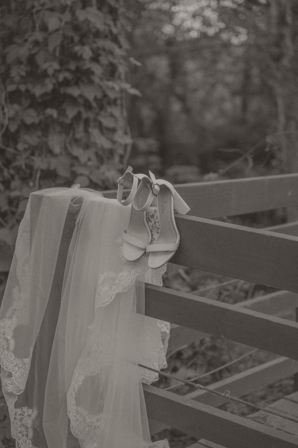 Black and white photo of a bridal veil and wedding shoes draped over a wooden fence outdoors.