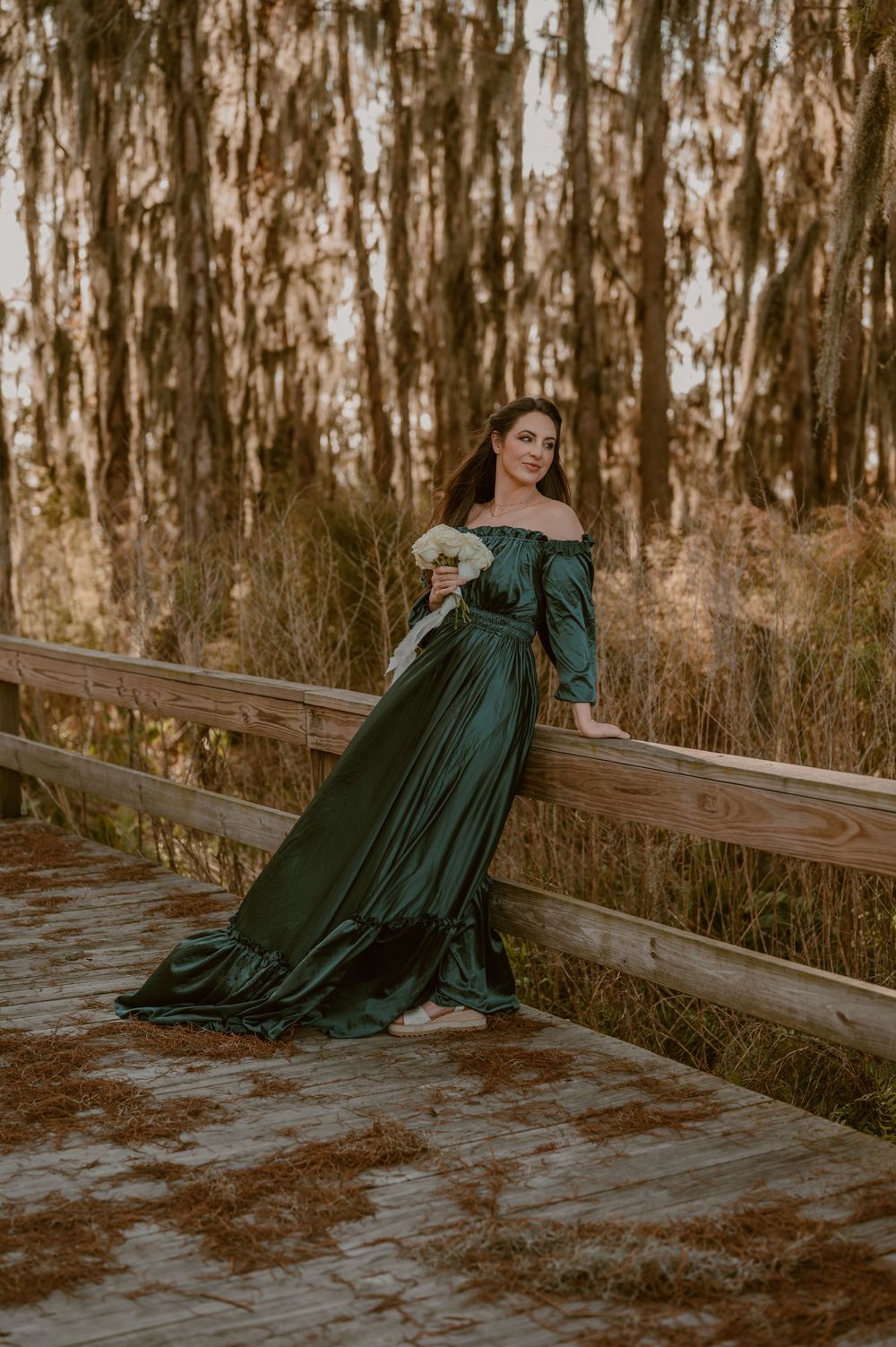 A long emerald green off-shoulder dress flows on wooden boardwalk surrounded by eucalyptus trees. photo by Samantha Campbell imagery