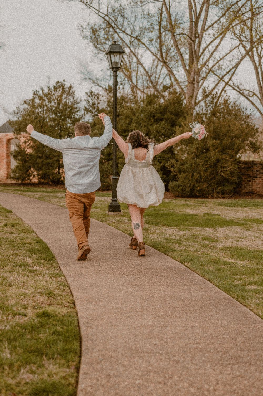 Couple joyfully skips down a path with their arms near a vintage streetlamp and trees after eloping. photo by Samantha Campbell imagery