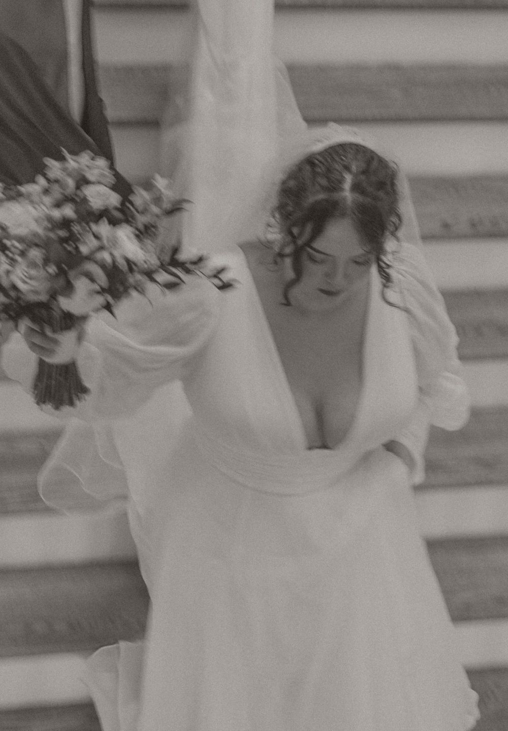 Bride in white wedding gown and veil holding floral bouquet on steps, black and white photo.