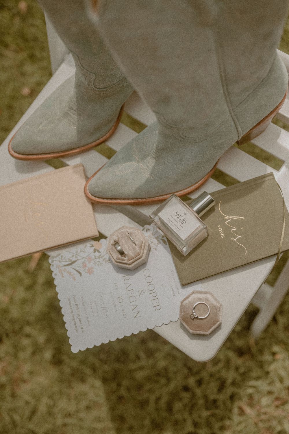 Rustic wedding flat lay with suede cowboy boots, floral invitation, perfume bottle, ring box, and elegant stationery.
