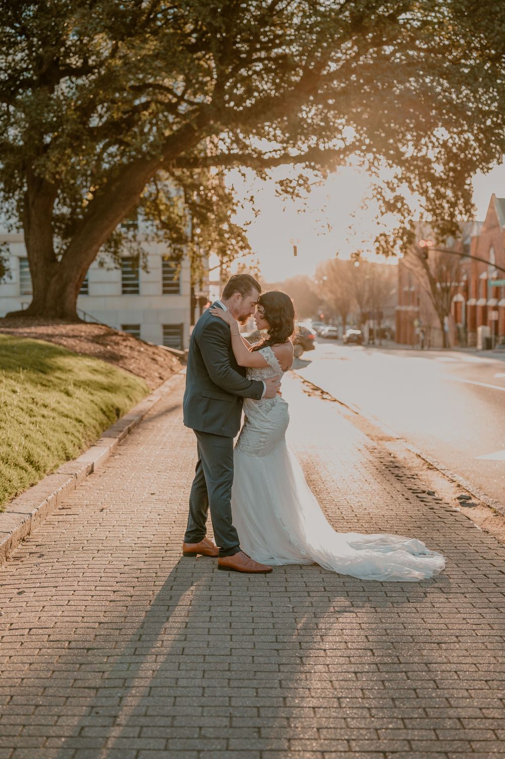 Newlyweds embrace on a brick pathway at sunset with golden light streaming through trees in the background.