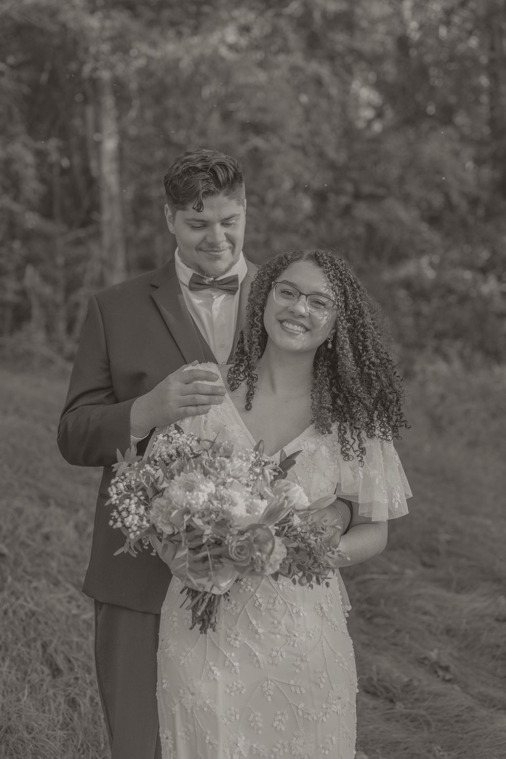 Black and white wedding photo of smiling bride holding bouquet with groom in bow tie outdoors.