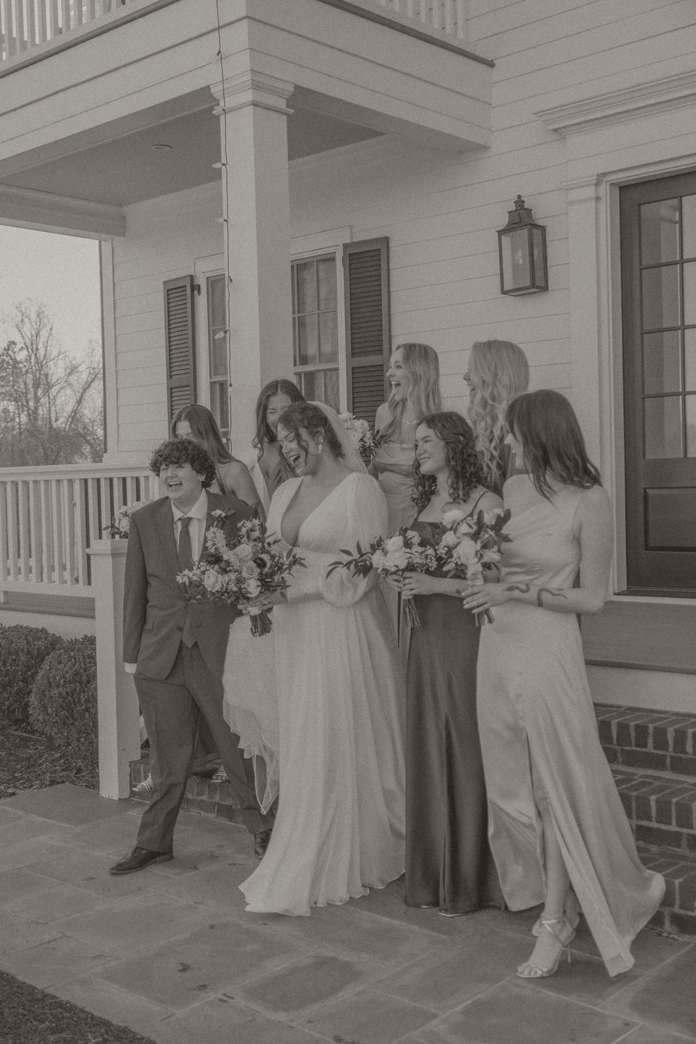 Black and white photo of a bride with bridesmaids holding bouquets outside a white Southern-style home.