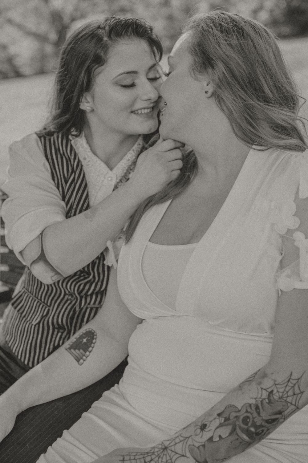 Black and white intimate portrait of two people sharing a tender romantic moment, one wearing a vest and one in white.
